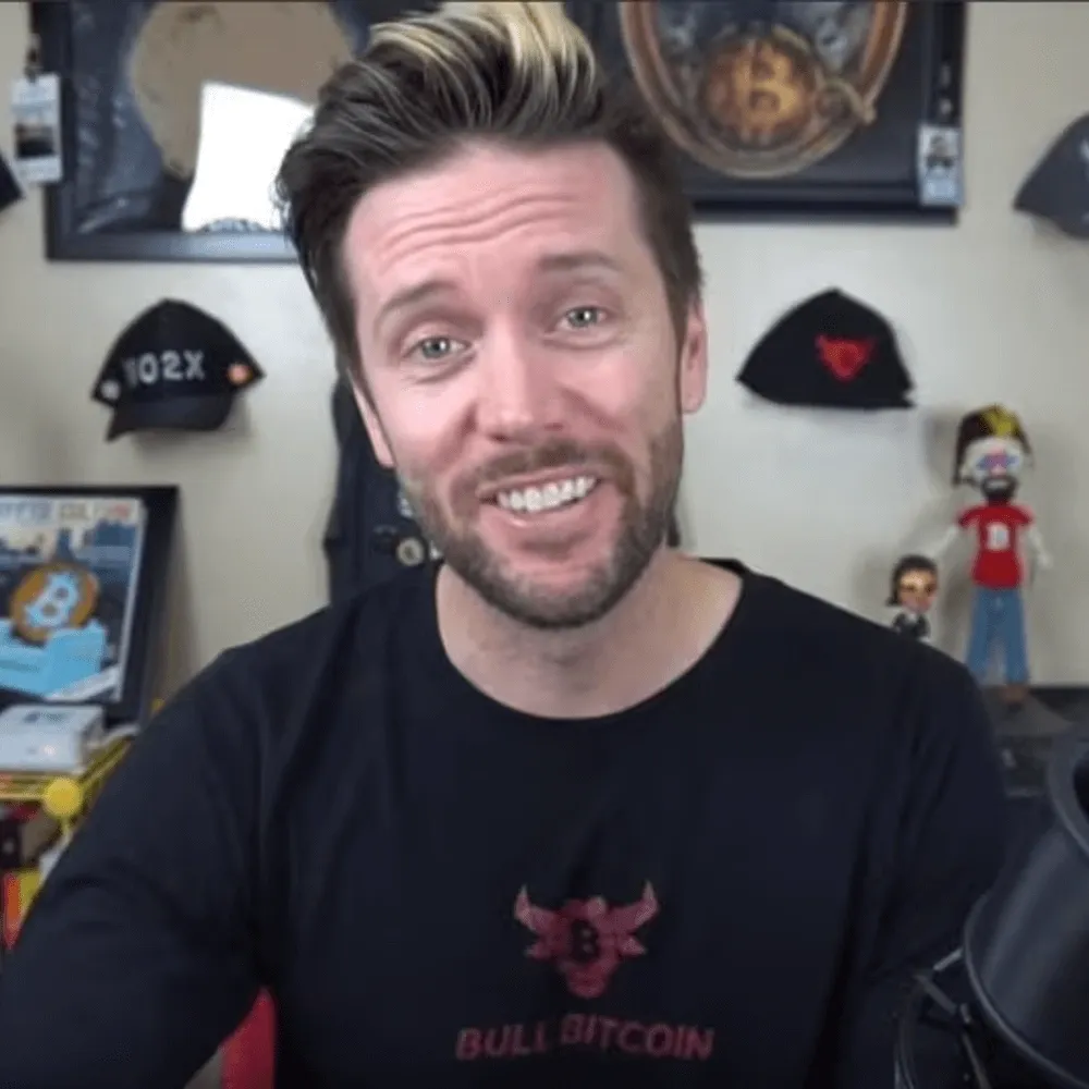 Man wearing a “Bull Bitcoin” shirt speaking on camera in a home office setup.
